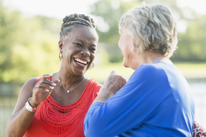 Two senior multi-ethnic woman, one Caucasian and the other African American, sitting in back yard by water enjoying each other's company. They are looking at each other, laughing.